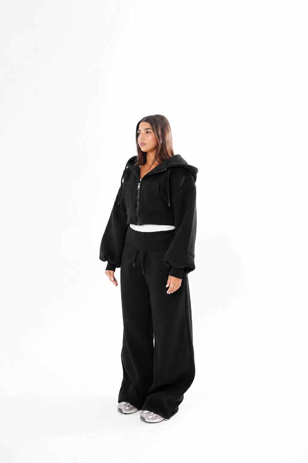 A woman modeling a black two-piece tracksuit consisting of a cropped zip-up hoodie with a hood and wide-leg sweatpants, standing against a solid white studio background.