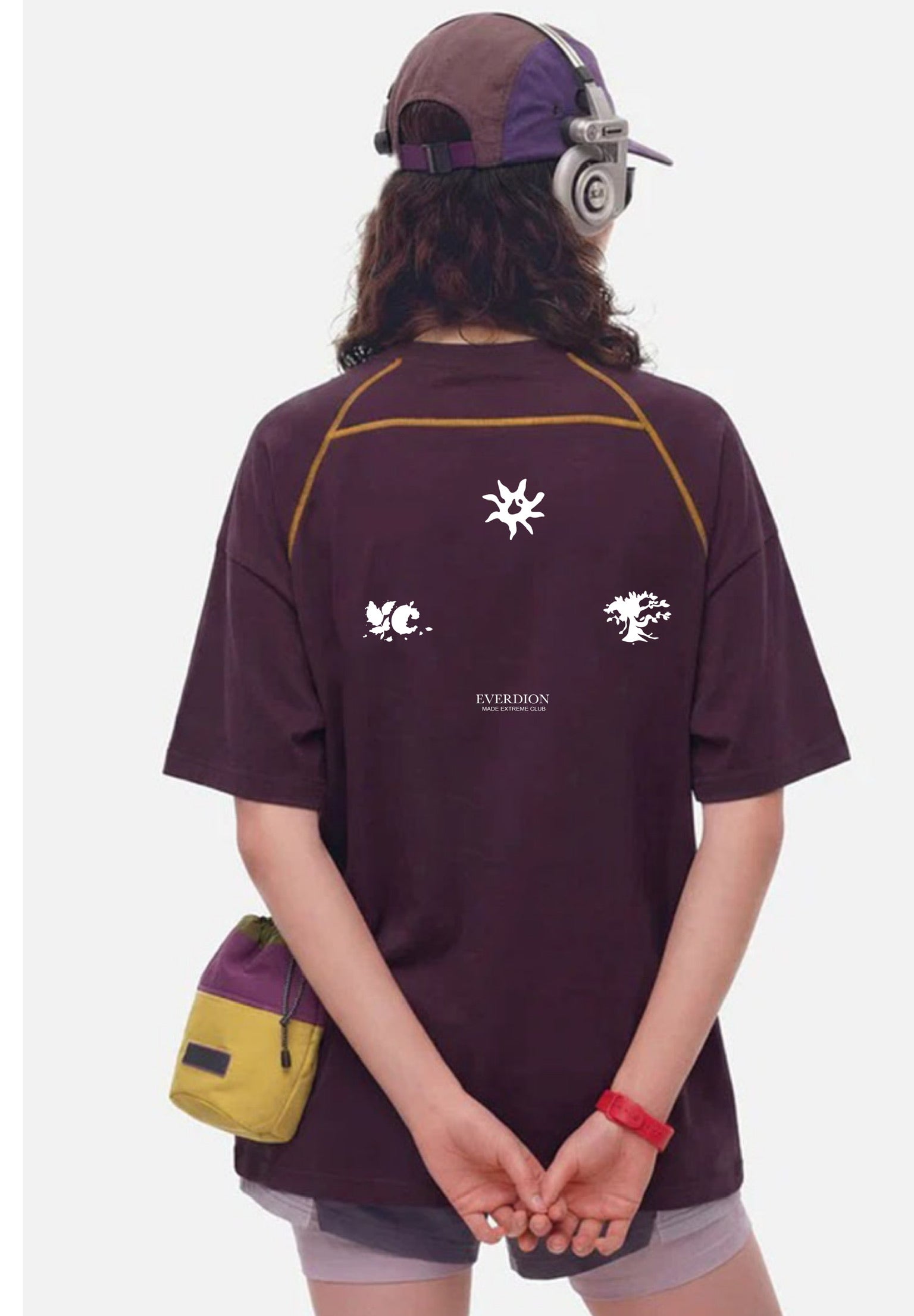 Person wearing a dark purple t-shirt with logos and text, holding a yellow bag on a white background