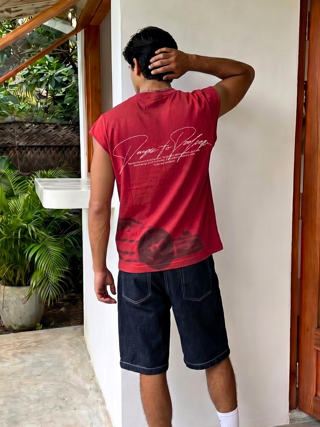 Person wearing a red sleeveless shirt with text and graphics, standing against a white wall.