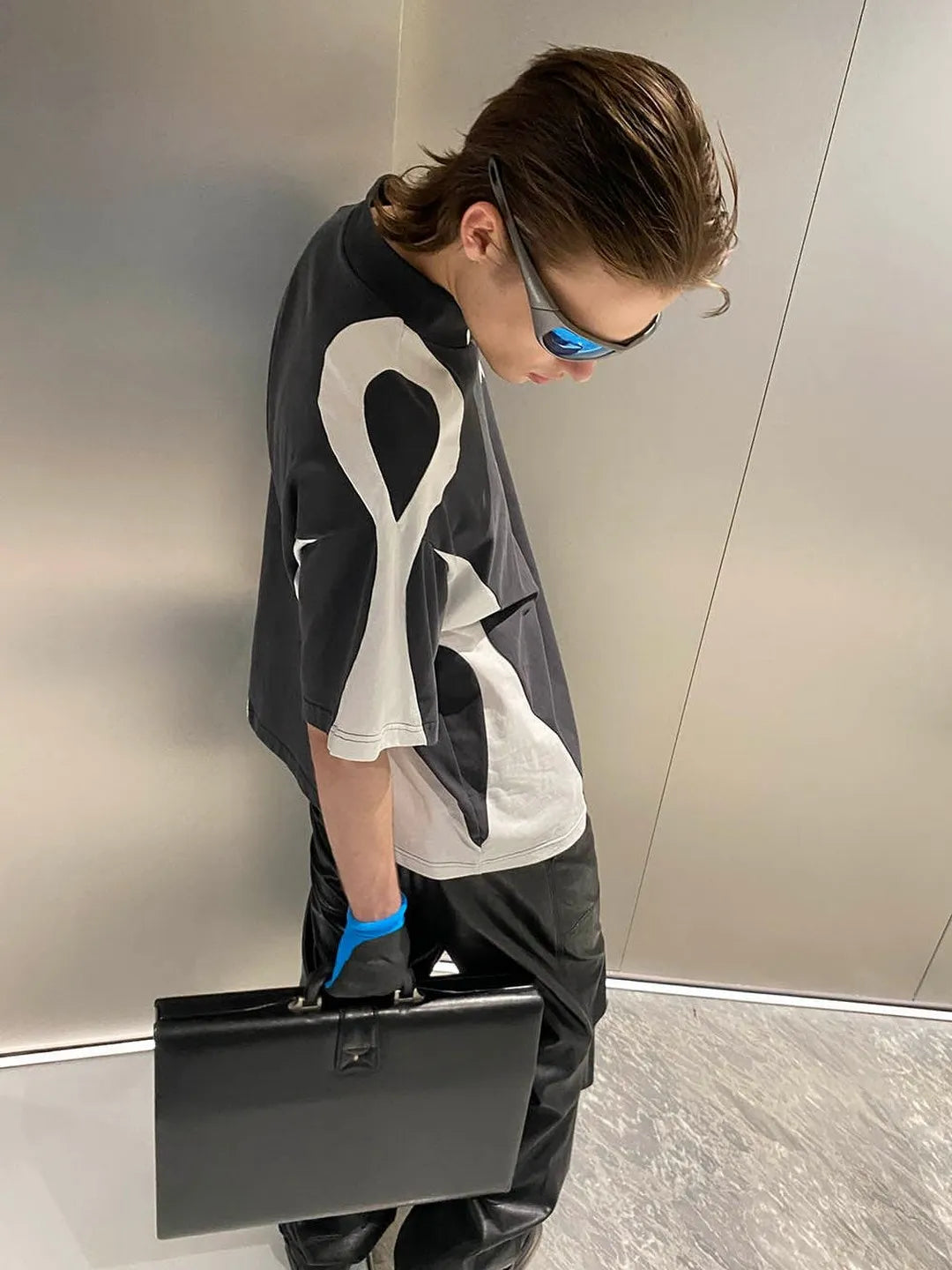 Person wearing a black and white outfit with a large logo, holding a black suitcase against a neutral background.