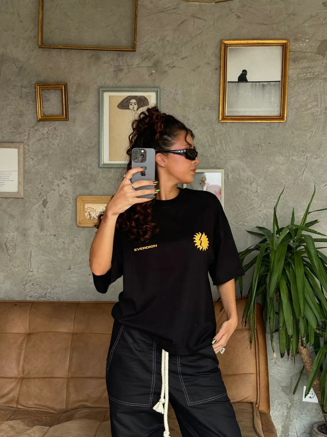 Person taking a mirror selfie wearing a black t-shirt with a yellow logo in an indoor setting.