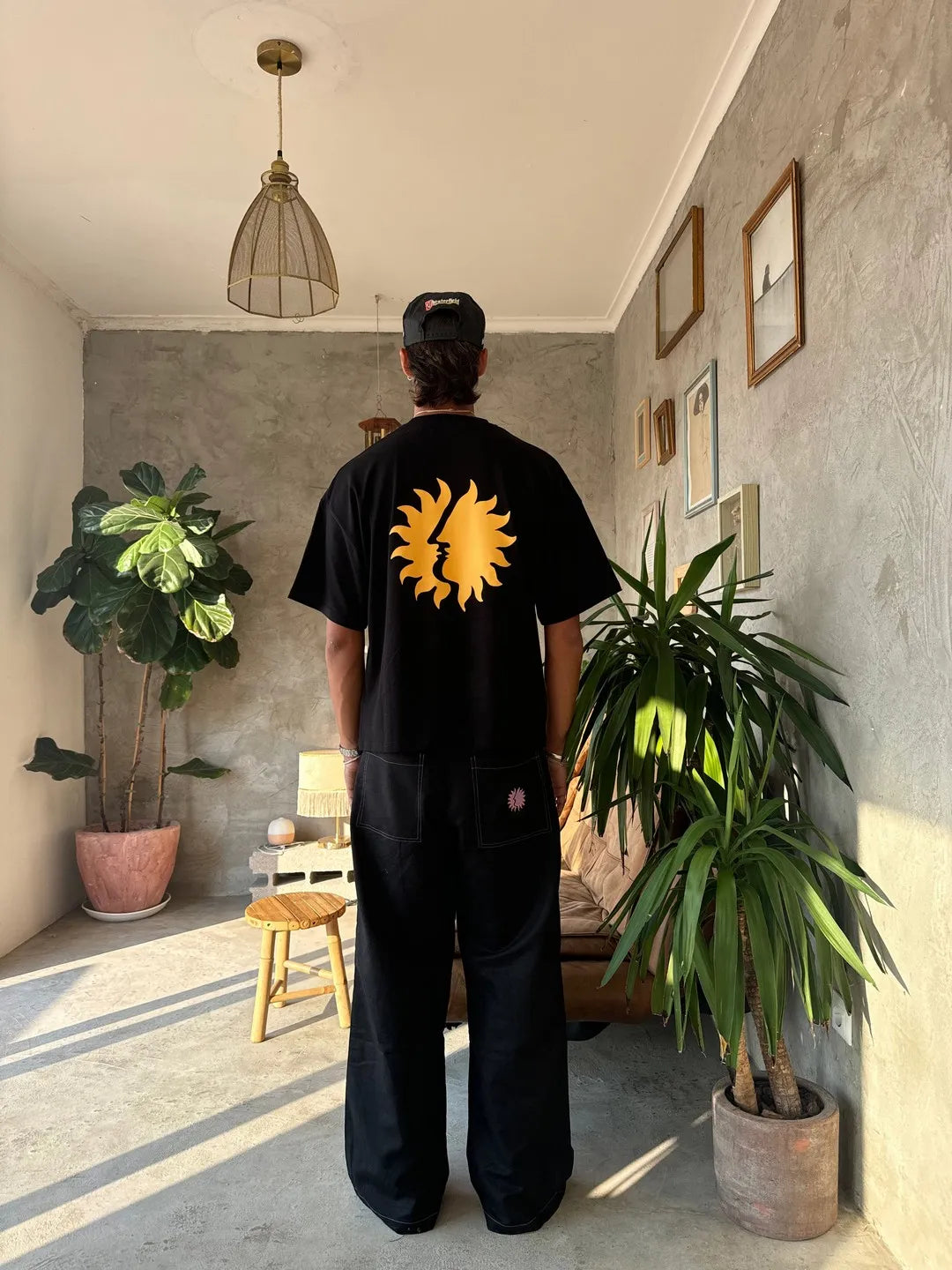 Person wearing a black t-shirt with a yellow sun design in a room with plants and a lamp.