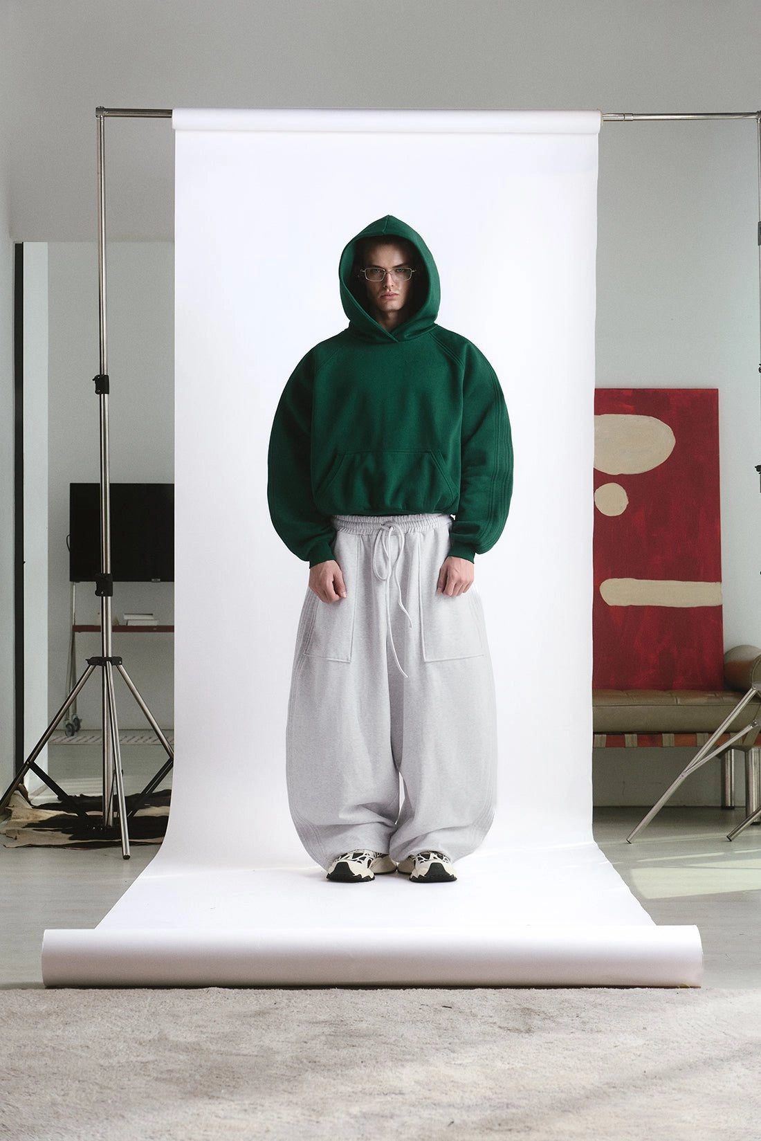 Person wearing a green hoodie and white pants standing in front of a white backdrop in a studio setting.