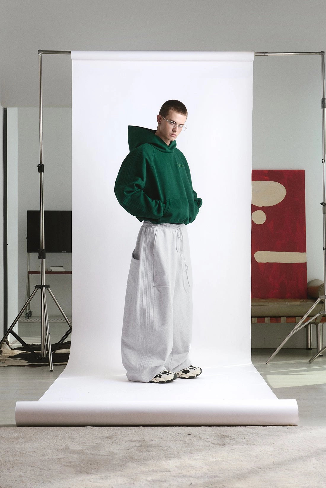 Person wearing a green hoodie and white pants standing in front of a white backdrop in a studio setting.