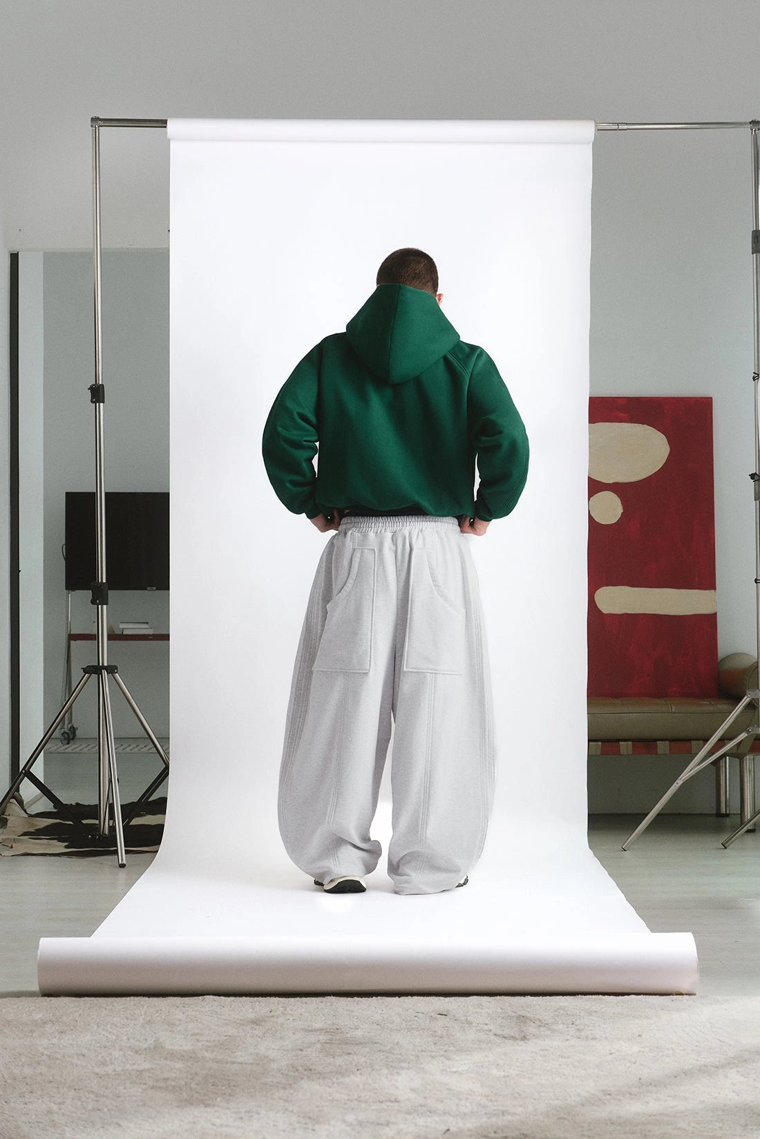 Person wearing a green hoodie and white pants standing in front of a large white backdrop in a studio setting.