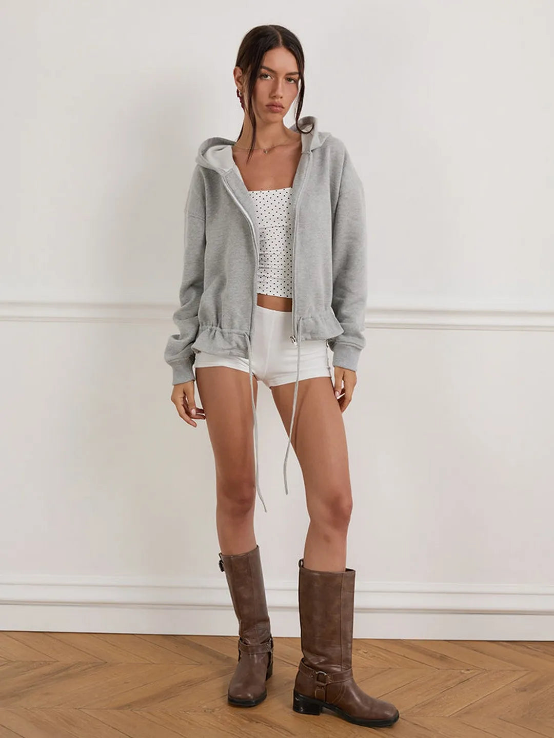 Woman wearing a gray hoodie, white patterned top, shorts, and brown boots against a white wall.