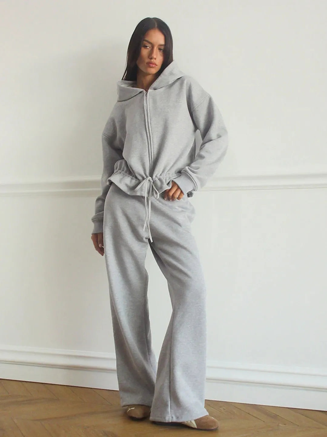Woman wearing a gray hoodie and pants against a white wall.