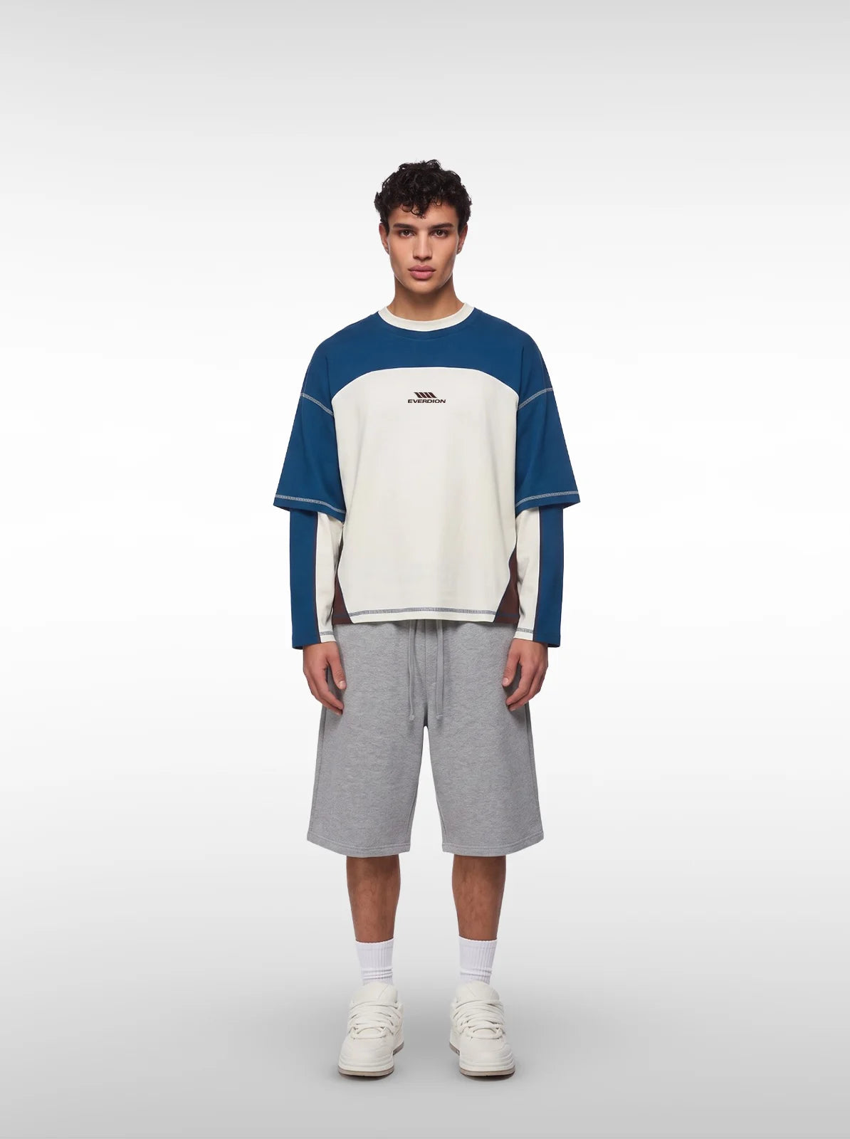 A male model wearing a color-blocked long-sleeve shirt with blue and off-white panels, grey shorts, white socks, and white sneakers.