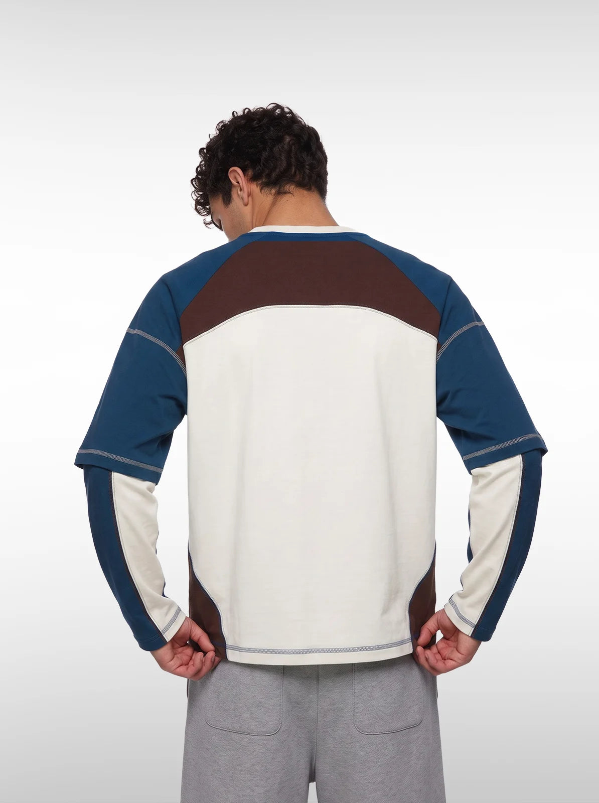 A man wearing a long-sleeve colorblock t-shirt with a cream body, blue sleeves, and brown shoulder panels, paired with gray sweatpants 