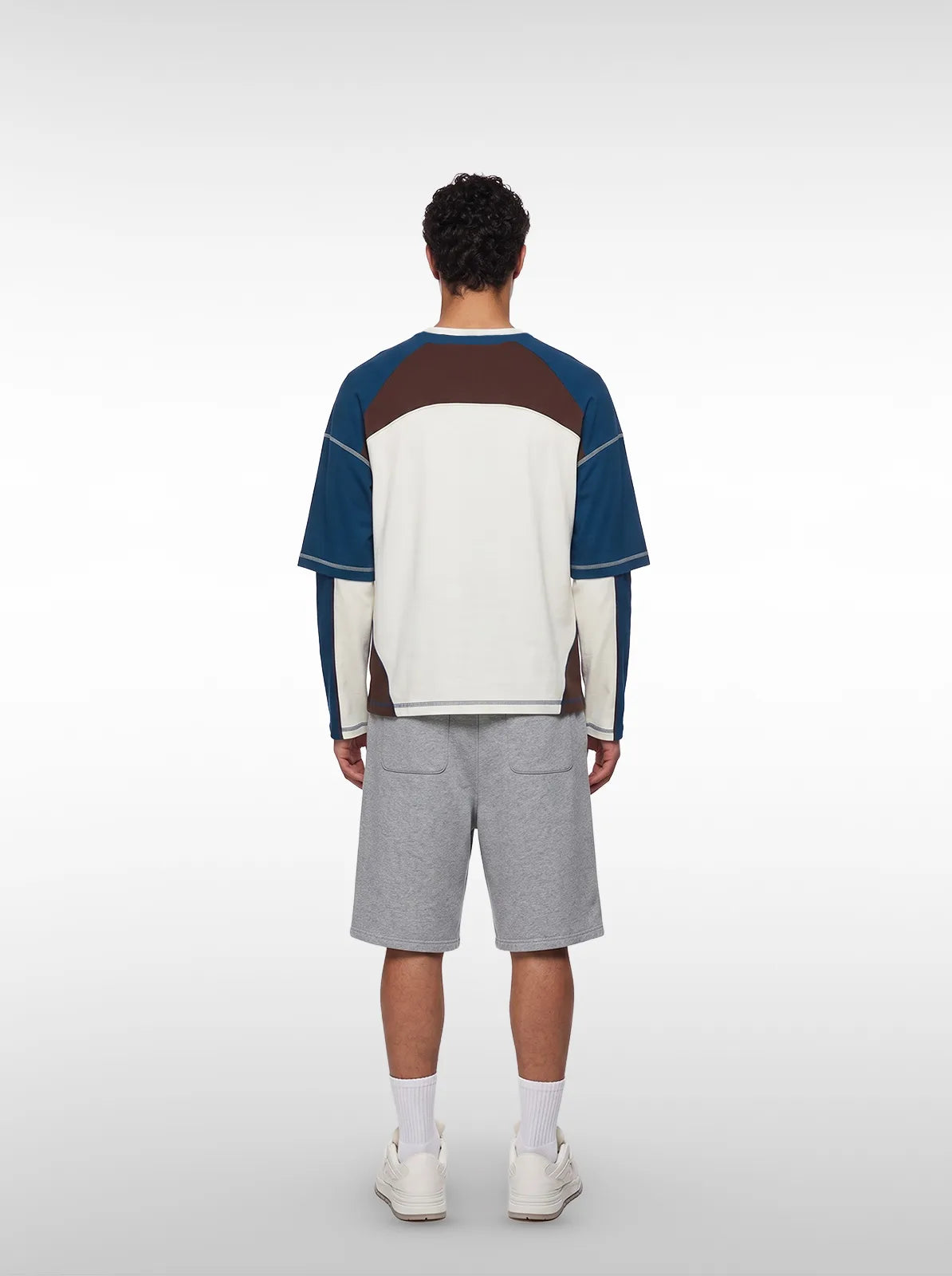A man wearing a color-blocked long-sleeve tshirt with blue, brown, and white panels, gray shorts, white socks, and white sneakers