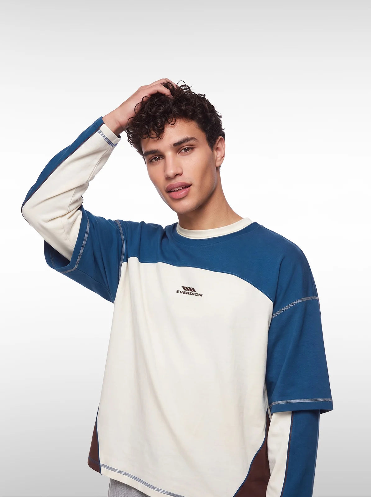 A young man with curly dark hair wearing a color-blocked long-sleeve shirt with blue and off-white panels, posed 