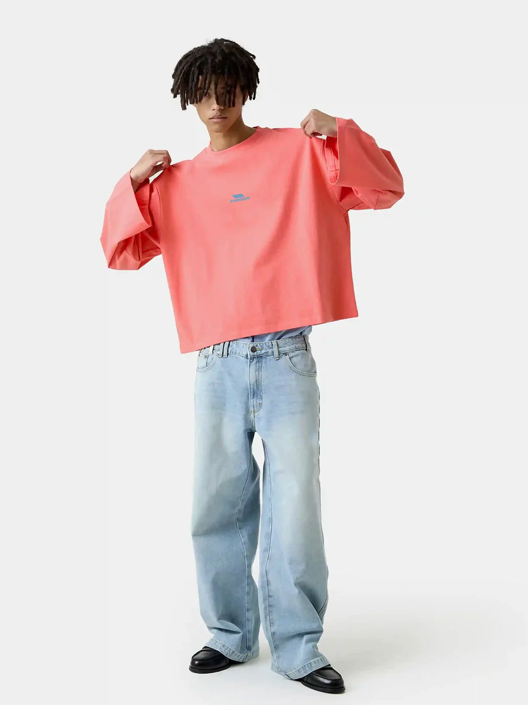 A model wearing a coral pink oversized crewneck tshirt with a small blue logo on the chest, paired with light-wash wide-leg jeans and black shoes, against a plain white studio background.