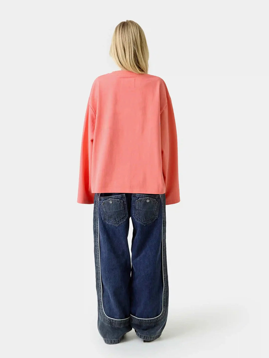 A model viewed from the back wearing a coral pink oversized long-sleeve top and dark blue wide-leg denim jeans with contrast stitching.