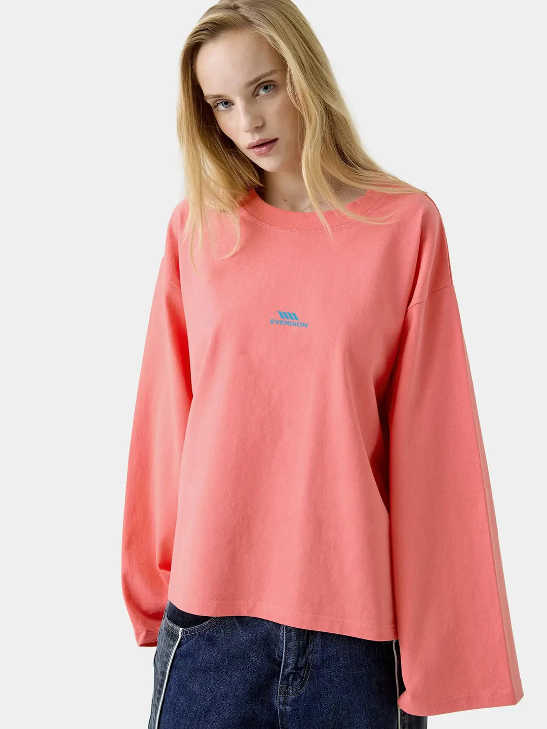 A model wearing a coral pink long-sleeve t-shirt with a small blue logo on the left chest, paired with dark blue jeans