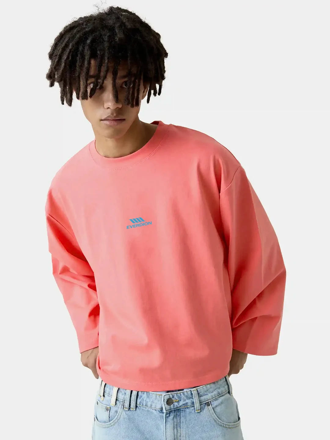 A model wearing a coral pink long-sleeve crewneck tshirt with a small blue graphic on the left chest, paired with light blue jeans