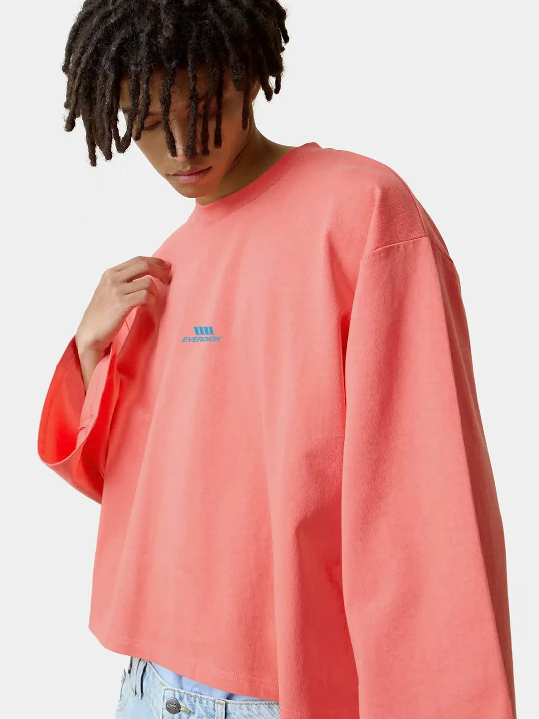 A model wearing a coral pink oversized long-sleeve t-shirt with a small blue logo on the left chest, paired with light blue jeans