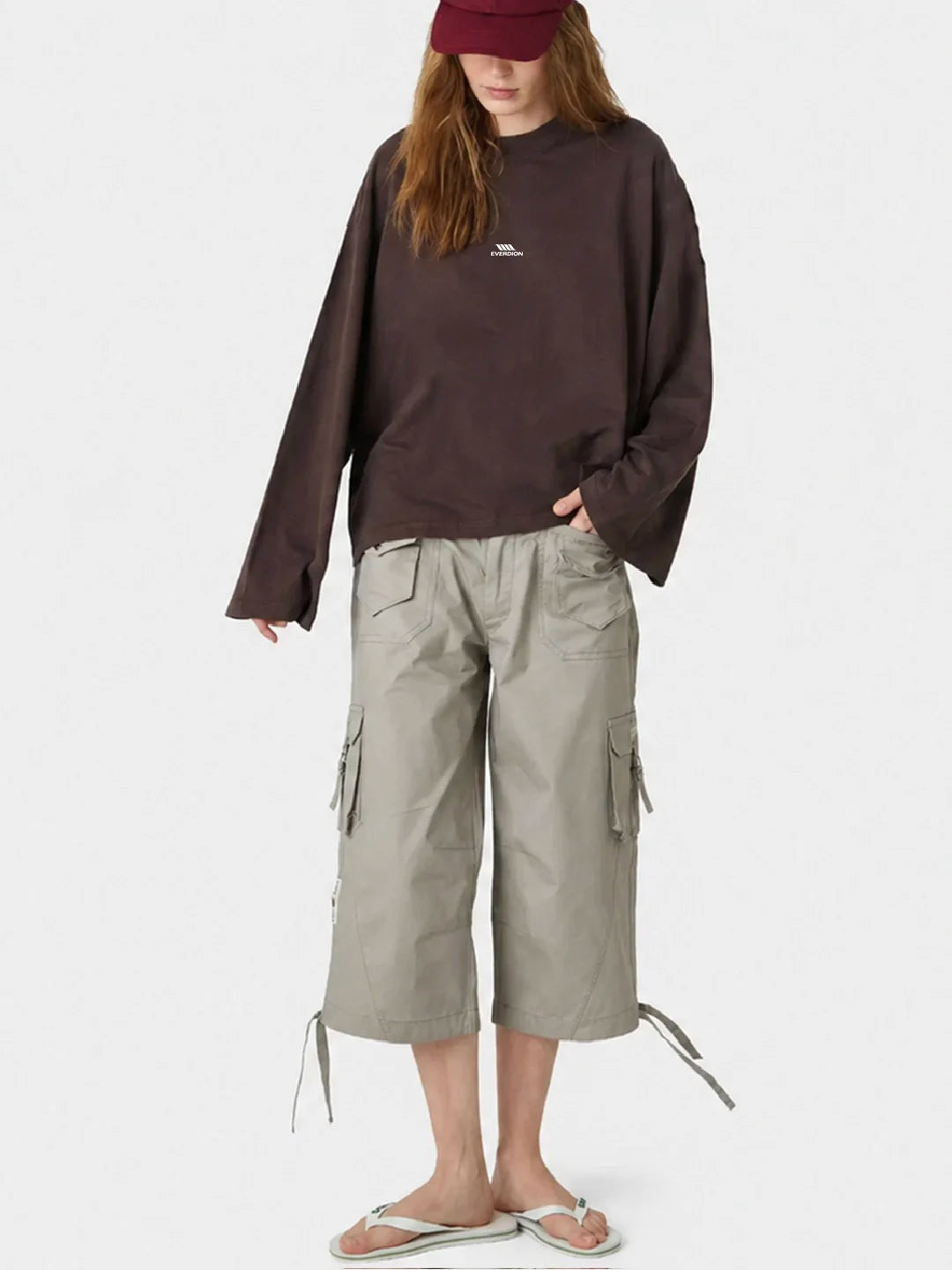 A model wearing an oversized dark brown tshirt, light grey cargo shorts, and a burgundy baseball cap, standing against a plain white studio background.