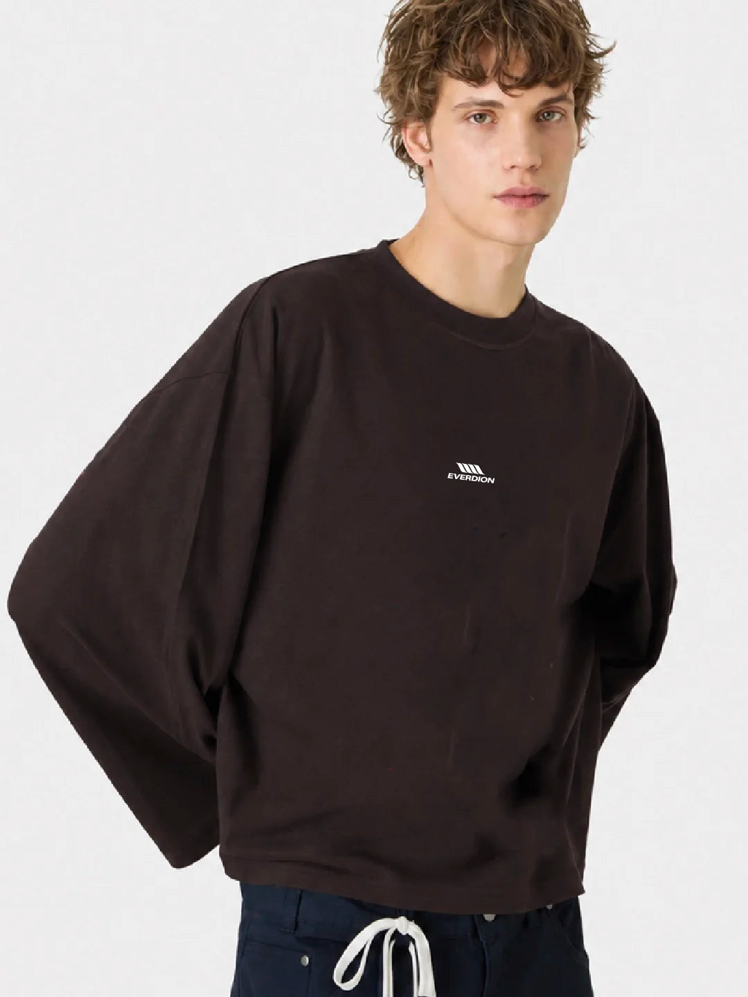 A young man wearing an oversized dark brown crewneck tshirt with a small white embroidered logo on the left chest, paired with dark pants
