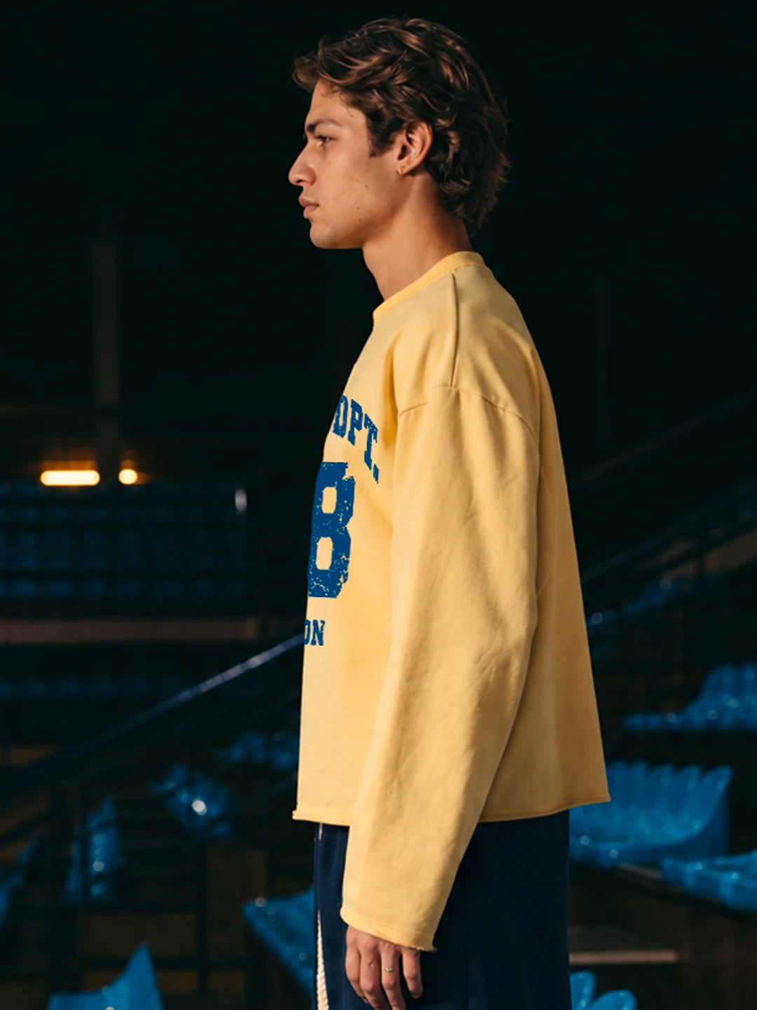 A man wearing a pale yellow long-sleeve tshirt with a large blue collegiate-style letter 'B' and partial text on the front, standing in a dark stadium or arena with blue seating in the background.