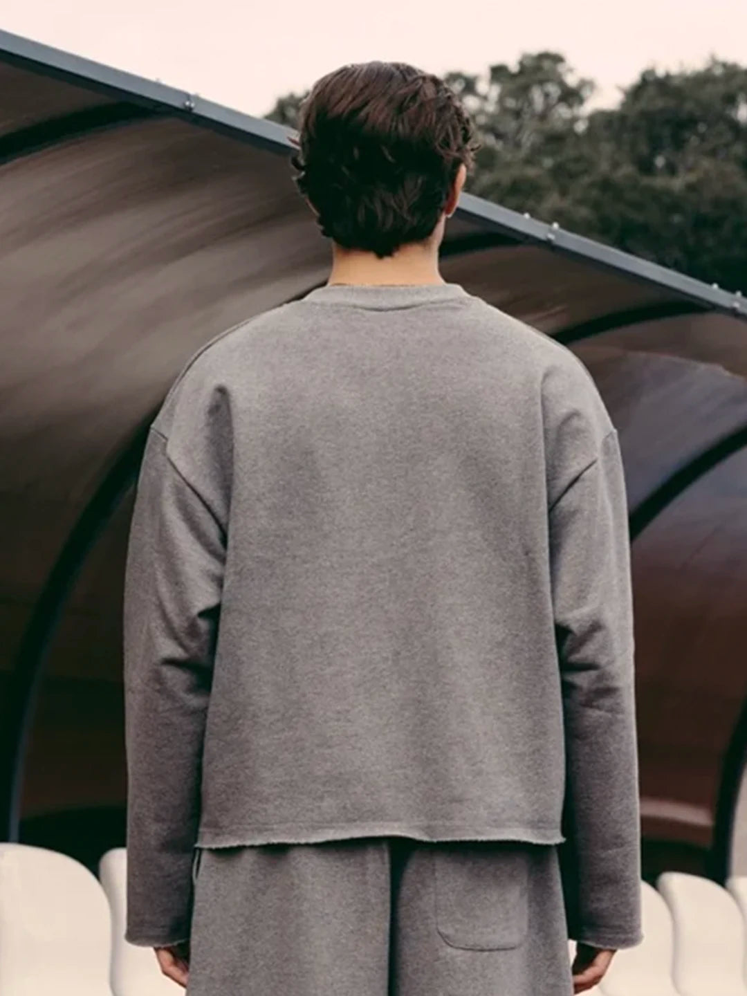 A person wearing a loose grey long-sleeve top and matching grey bottoms, standing under a curved roof structure outdoors.