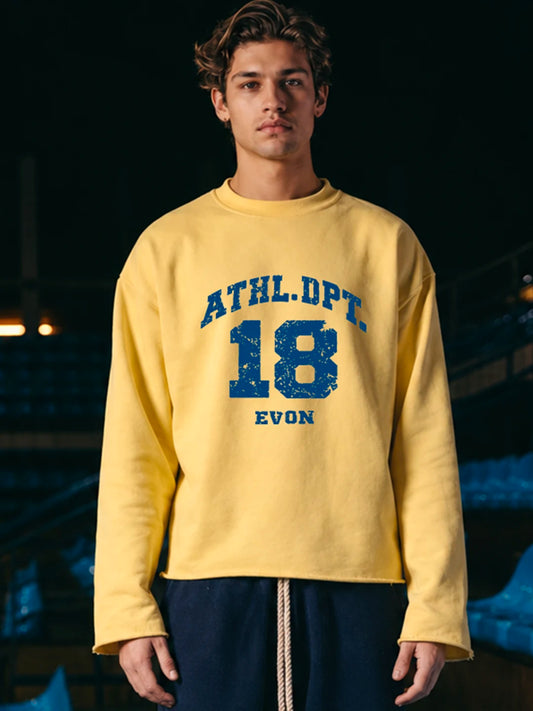 A young man wearing a yellow crewneck tshirt with a blue distressed collegiate-style graphic reading 'ATHL.DPT.
