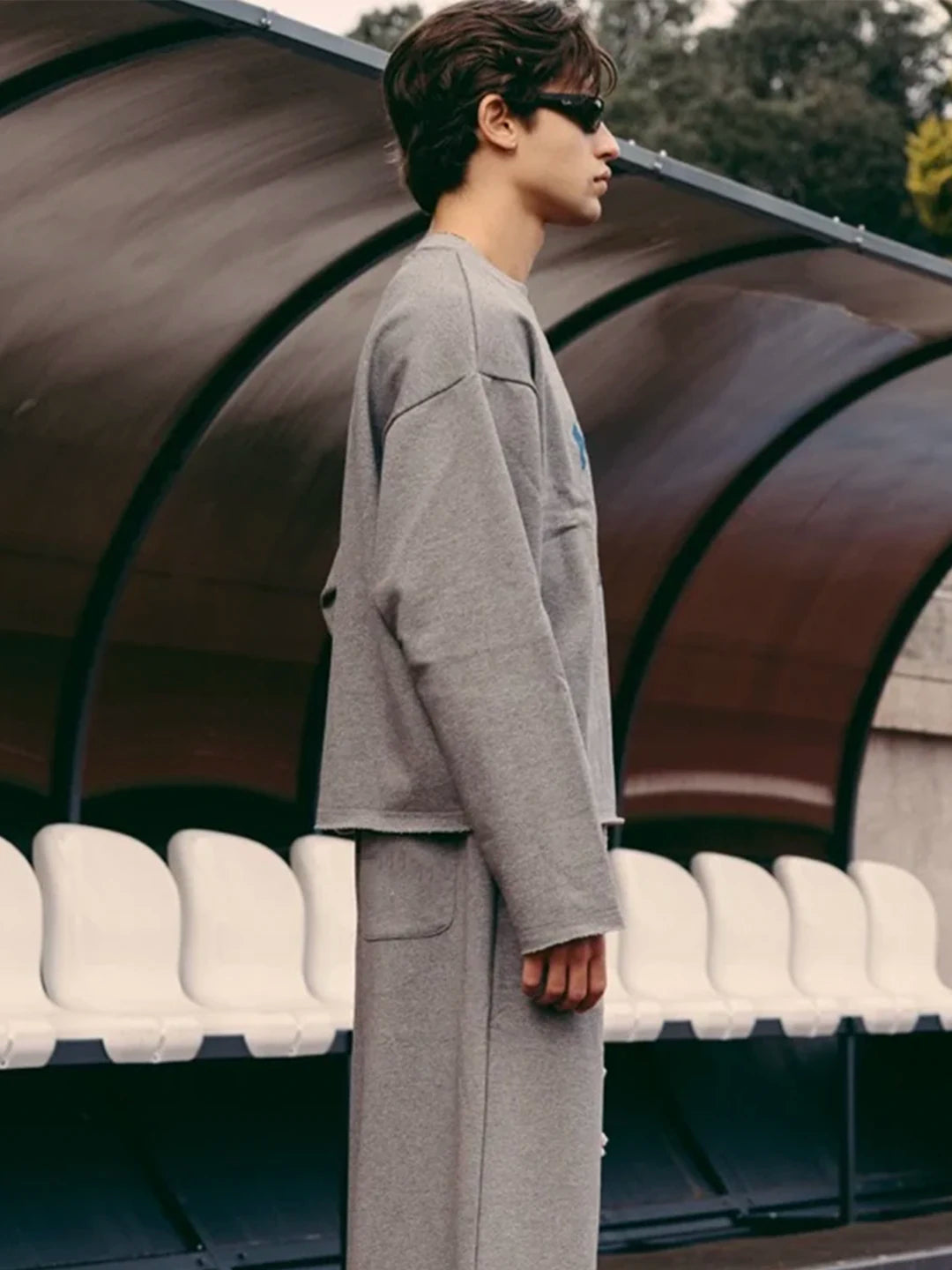 A man wearing a light grey oversized long-sleeve t-shirt and matching wide-leg trousers, standing in profile in front of stadium seating under a curved roof.