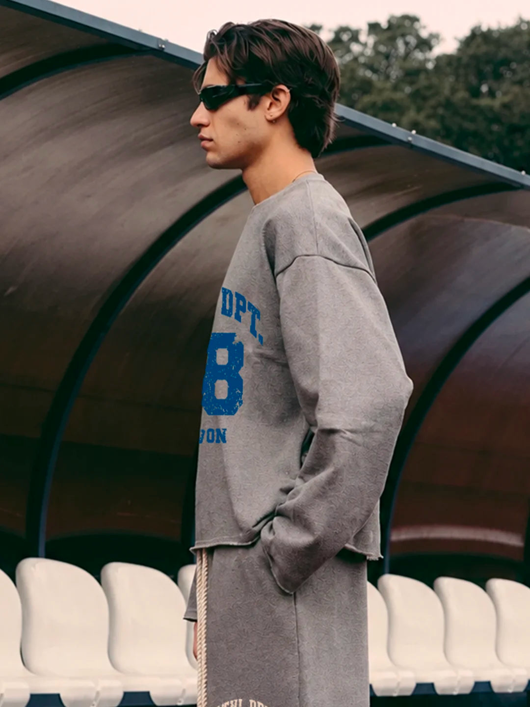 A man in profile wearing a grey oversized tshirt with blue collegiate-style lettering and a matching gry sweatpants standing in front of a covered outdoor seating area.