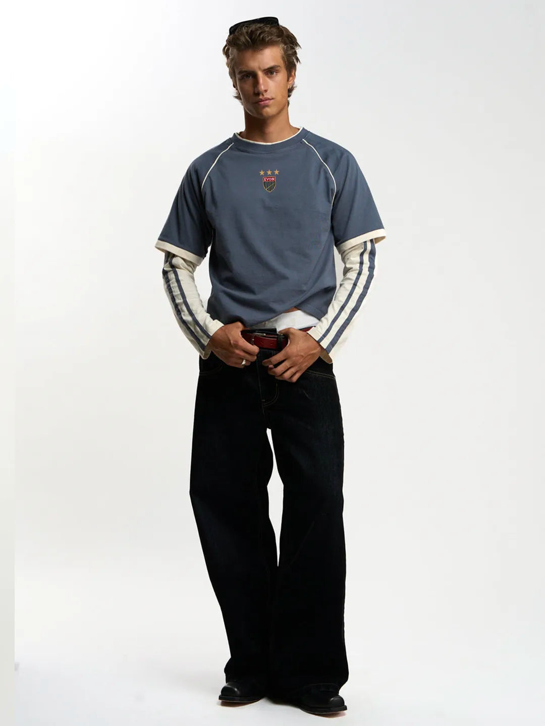 A man modeling a slate blue long-sleeve shirt with white raglan sleeves and a small red and yellow chest graphic, paired with black jeans against a white studio background.