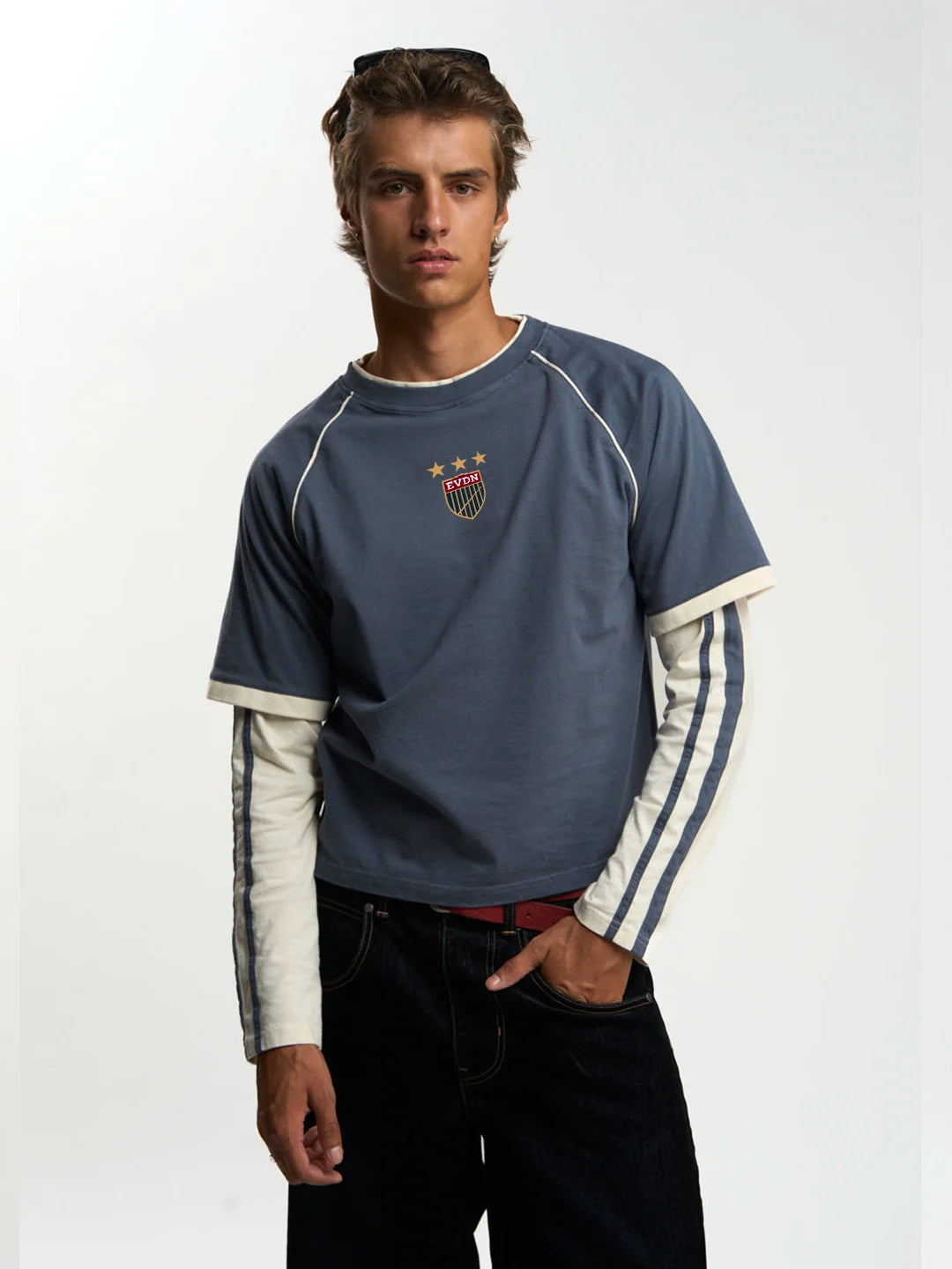 A male wearing a blue and white raglan-style long-sleeve t-shirt with a small crest on the chest, paired with dark jeans against a plain white studio background.