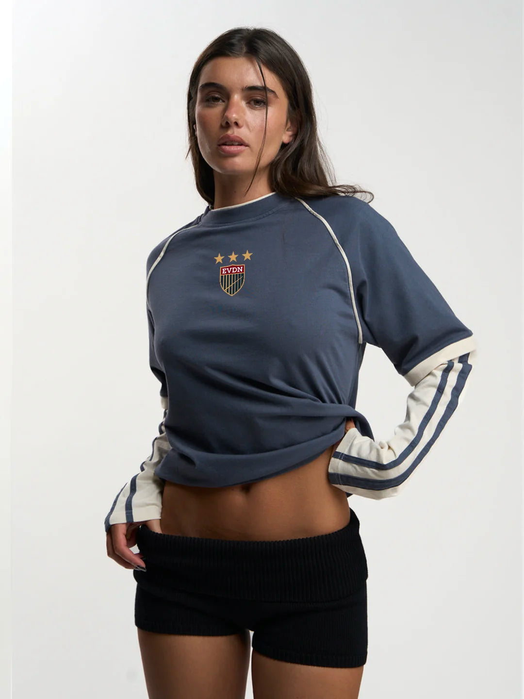A woman modeling a navy blue cropped long-sleeve sweatshirt with white contrast stitching, a small shield crest on the chest, and white stripes on the sleeves, paired with black shorts against a plain light background.