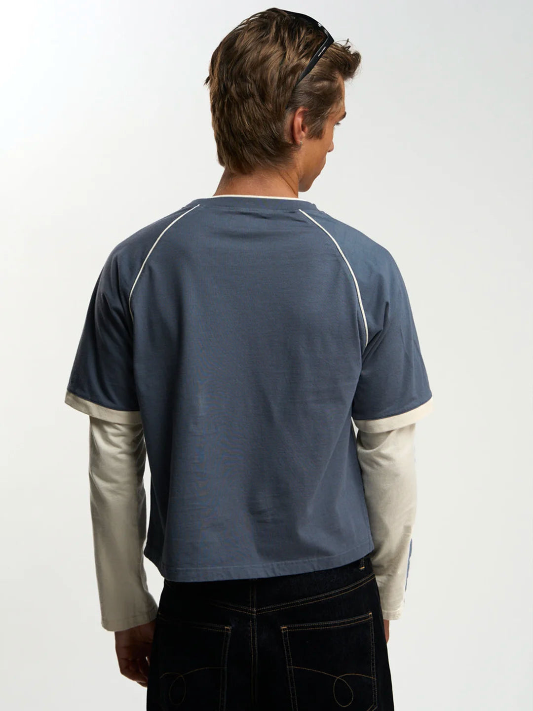 A man viewed from behind wearing a slate blue short-sleeve t-shirt with contrasting cream-colored raglan sleeves and collar trim, layered over a cream long-sleeve shirt and paired with dark jeans.