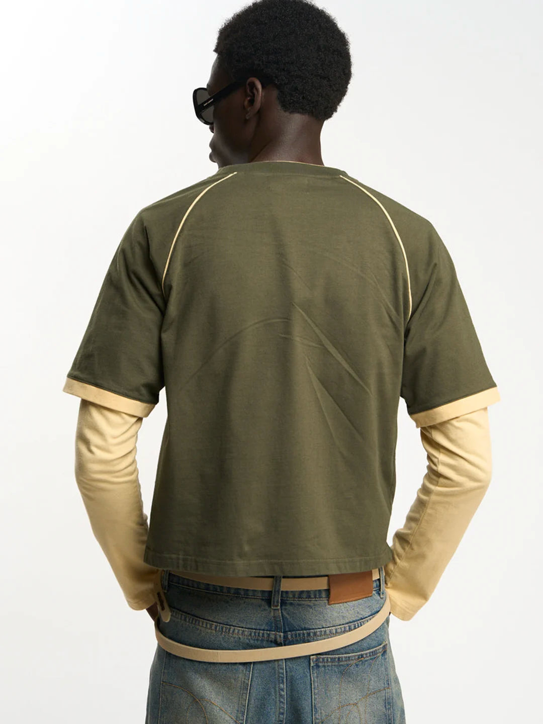 A man wearing an olive green short-sleeve t-shirt layered over a beige long-sleeve shirt, paired with blue jeans and black sunglasses, against a plain white background.