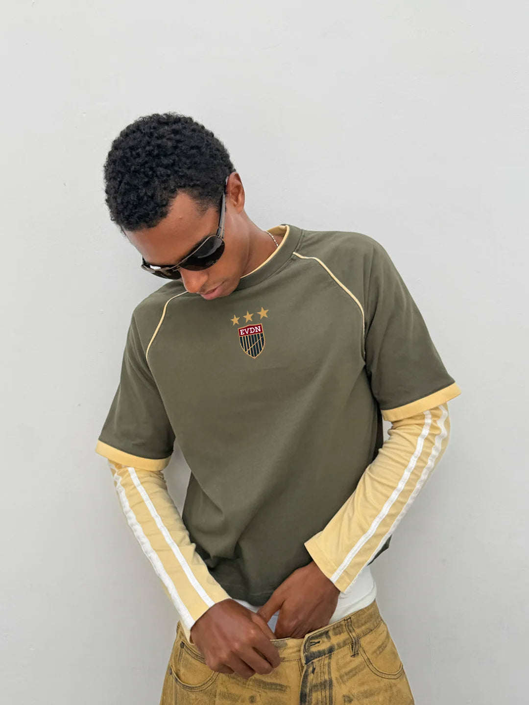 A man wearing an olive green short-sleeve t-shirt with yellow and white raglan sleeves, a small shield crest on the chest, and a white long-sleeve shirt underneath, paired with tan jeans 