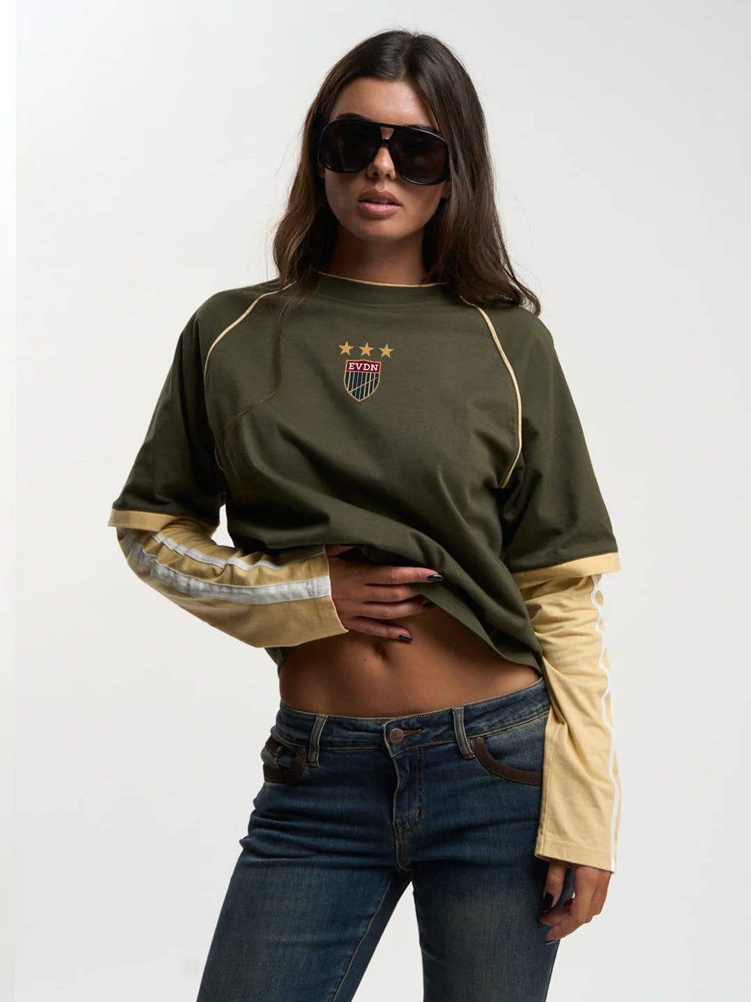A woman modeling a cropped long-sleeve t-shirt with an olive green body, beige sleeves, and a small shield crest on the chest, paired with blue jeans against a plain light background.