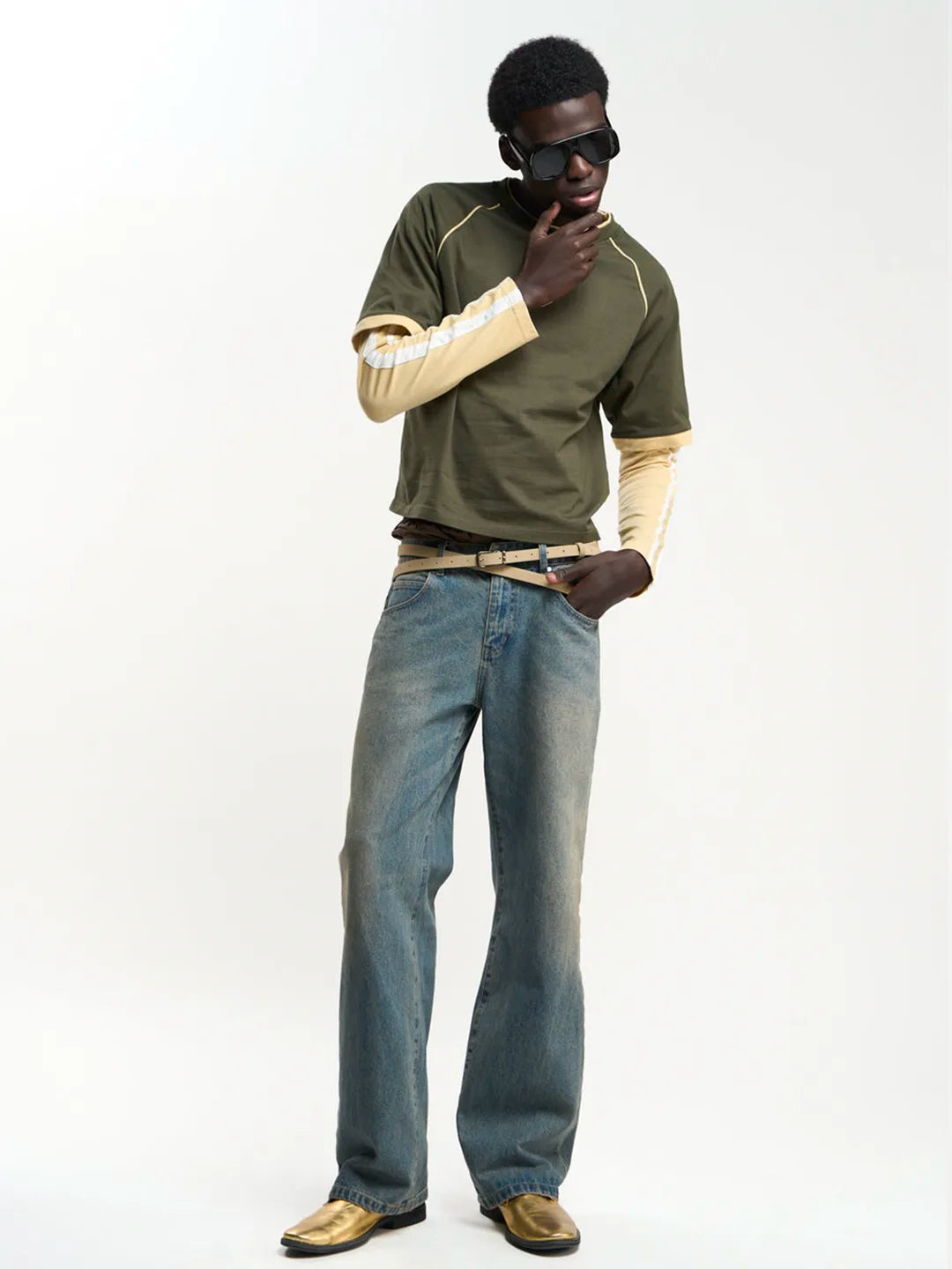 A man modeling a green short-sleeve t-shirt with beige sleeves, paired with blue jeans, a tan belt, and gold loafers, against a plain white studio background.