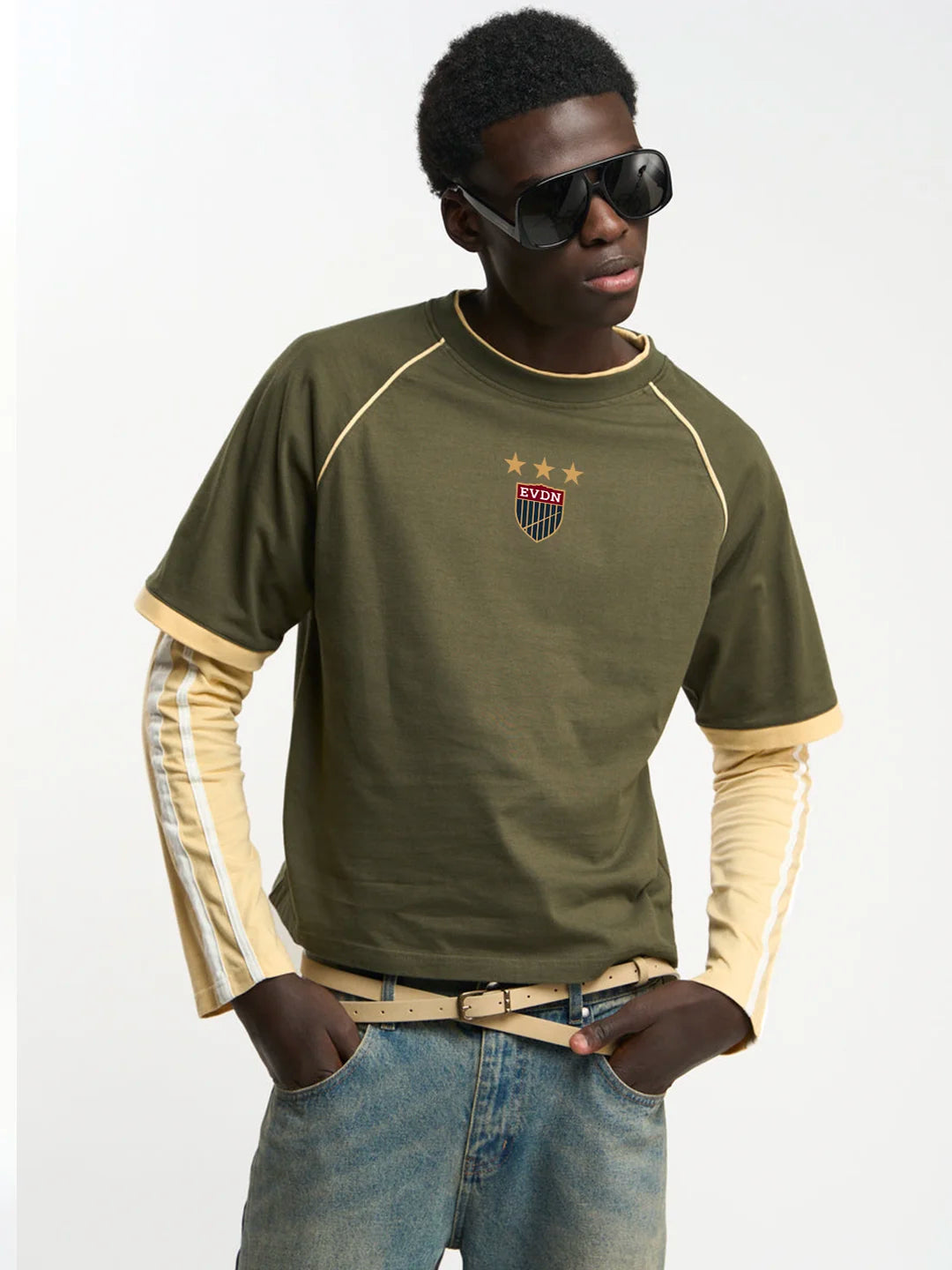 A man wearing an olive green short-sleeve t-shirt with beige contrast stitching and a small shield crest on the chest, layered over a beige long-sleeve shirt, paired with blue jeans and black sunglasses, against a plain white background.