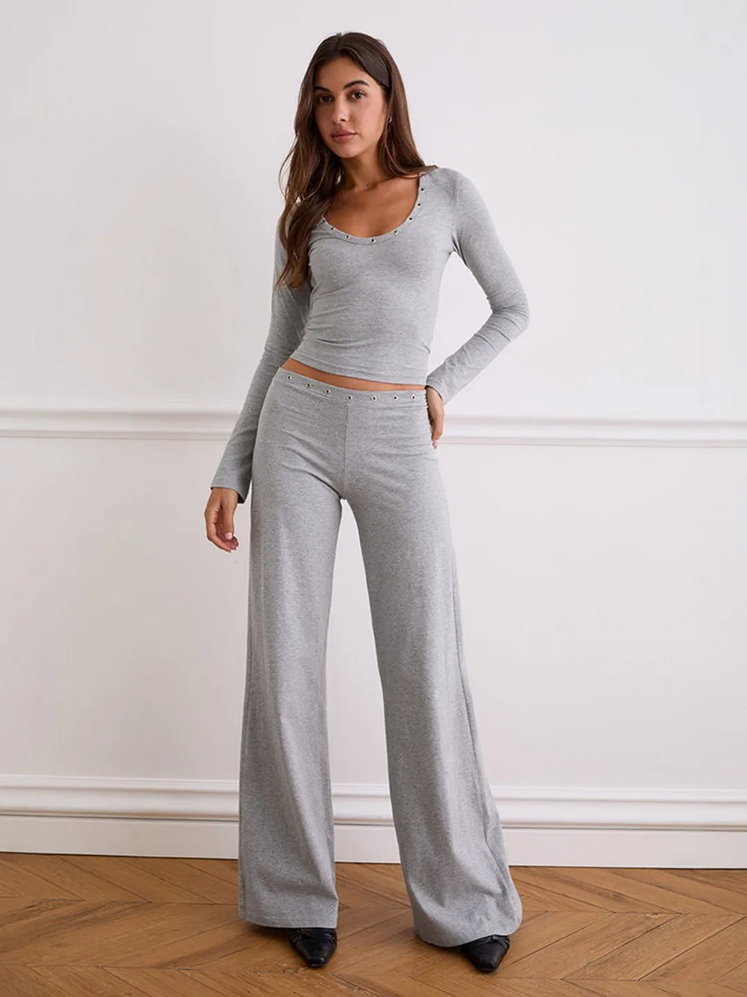 Woman wearing a gray long-sleeve top and wide-leg pants against a white wall.