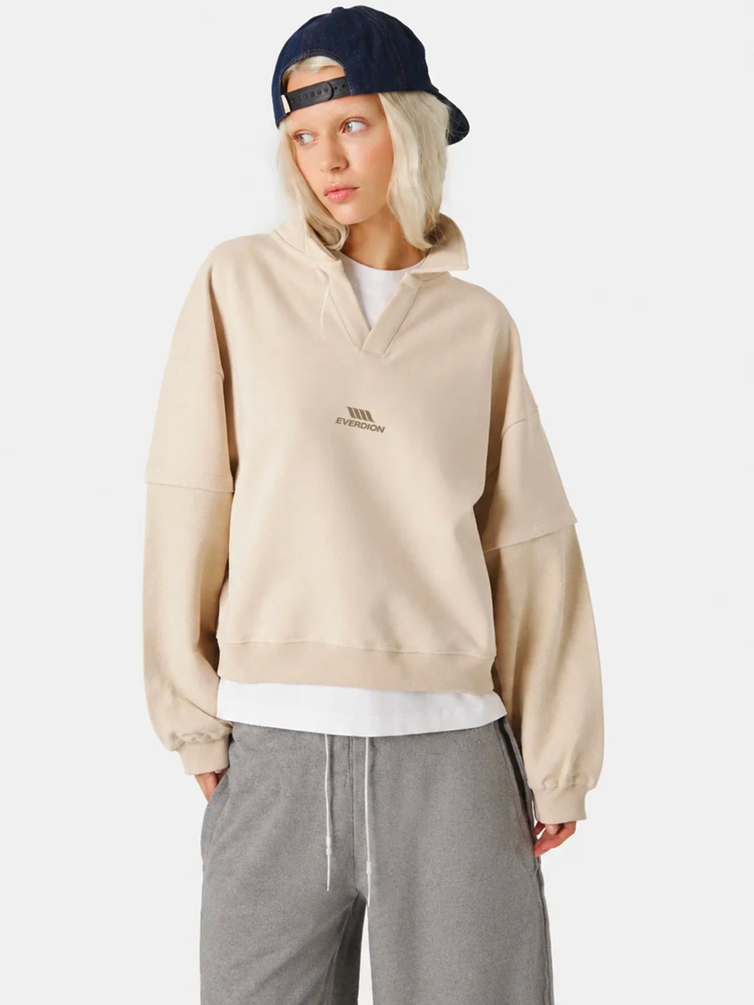 A model wearing a beige polo t-shirt with a small embroidered logo on the chest, styled with grey sweatpants, a white undershirt, and a navy blue baseball cap 
