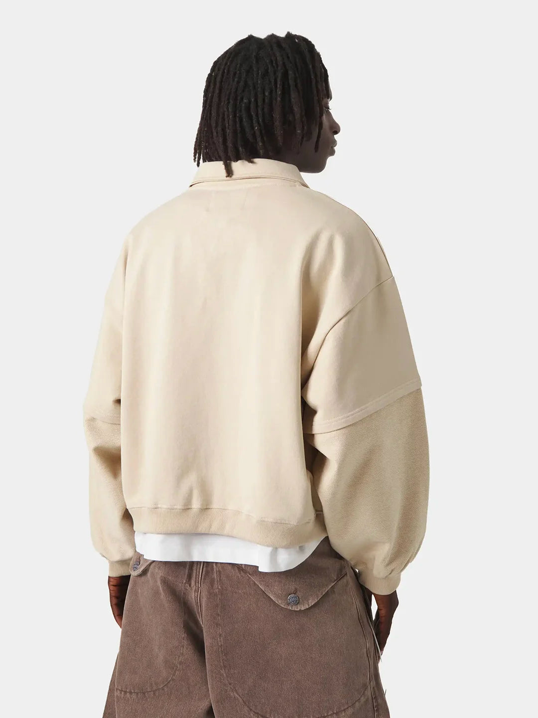 A person wearing a light beige polo t-shirt over a white shirt and brown corduroy pants, against a plain light gray studio background.