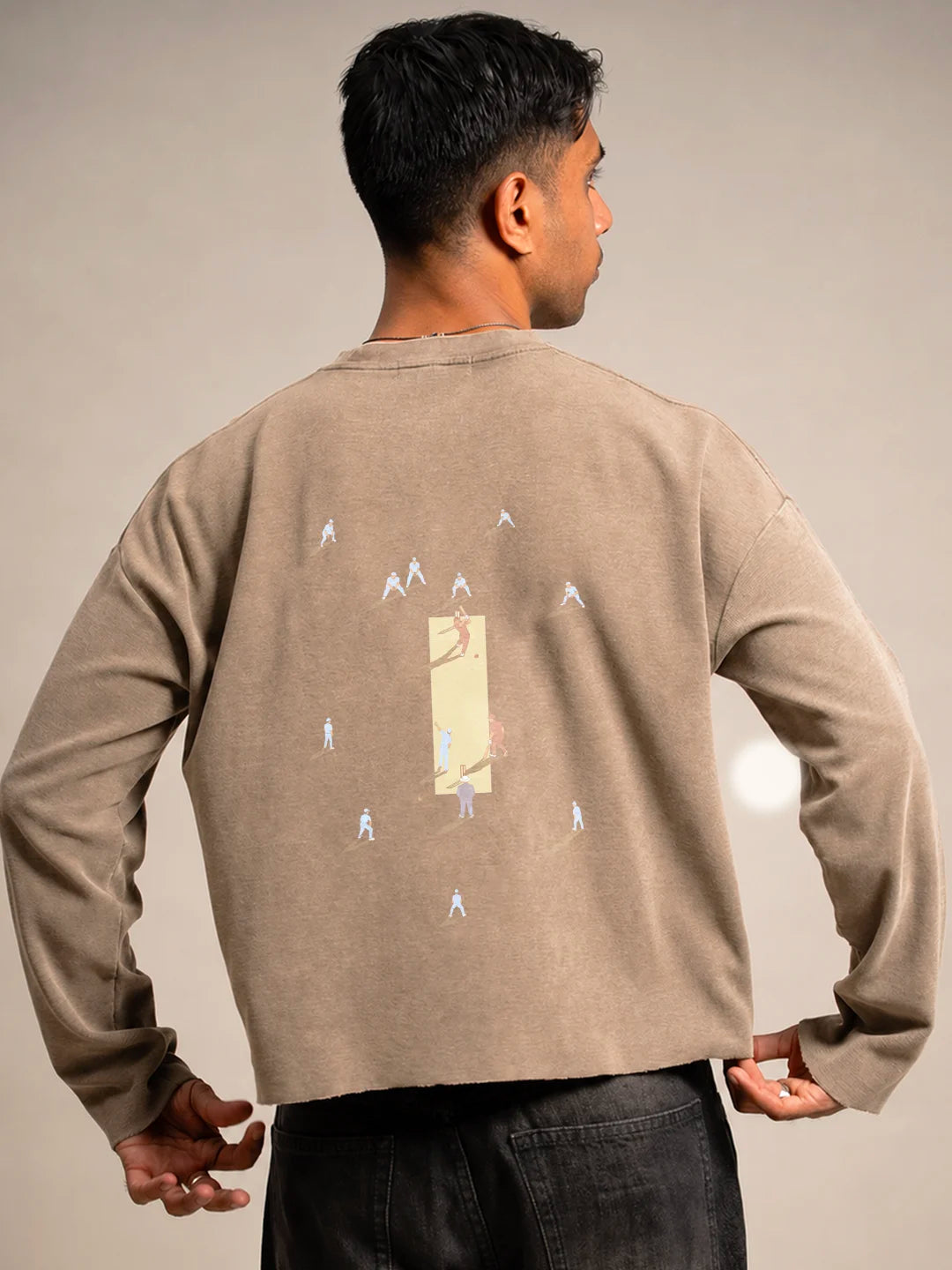 A man viewed from behind wearing a beige crewneck t-shirt with a graphic print on the back, paired with black jeans, against a plain beige studio background.
