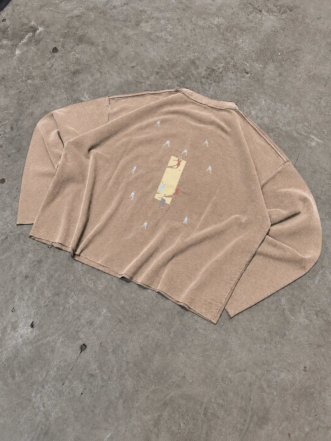 A tan long-sleeve knit t-shirt with a small central graphic print, laid flat on a textured grey concrete surface.