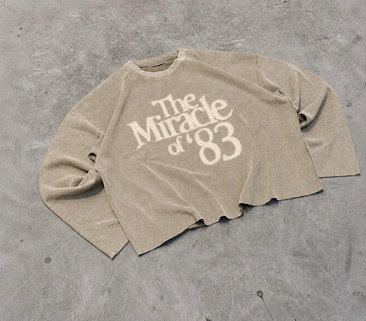 A beige cropped long-sleeve t-shirt with a distressed cream graphic reading 'The Miracle of '83' laid flat on a grey concrete floor.