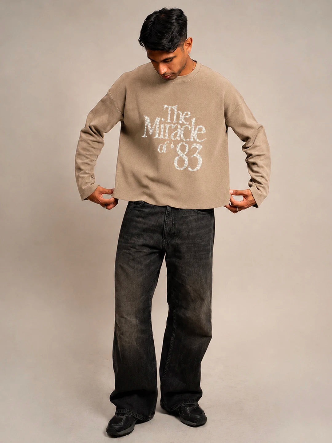 A man wearing a beige long-sleeve t-shirt with 'The Miracle of 83' printed in white on the front, paired with dark grey jeans and black shoes, standing against a neutral beige background.