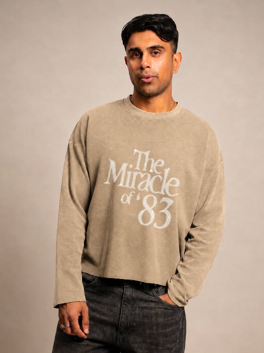A man wearing a beige crewneck t-shirt with a distressed white graphic reading 'The Miracle of 83' on the front, paired with dark grey jeans, against a neutral beige studio background.