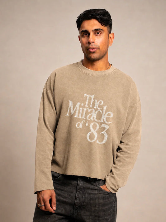 A man wearing a beige crewneck t-shirt with a distressed white graphic reading 'The Miracle of 83' on the front, paired with dark grey jeans, against a neutral beige studio background.