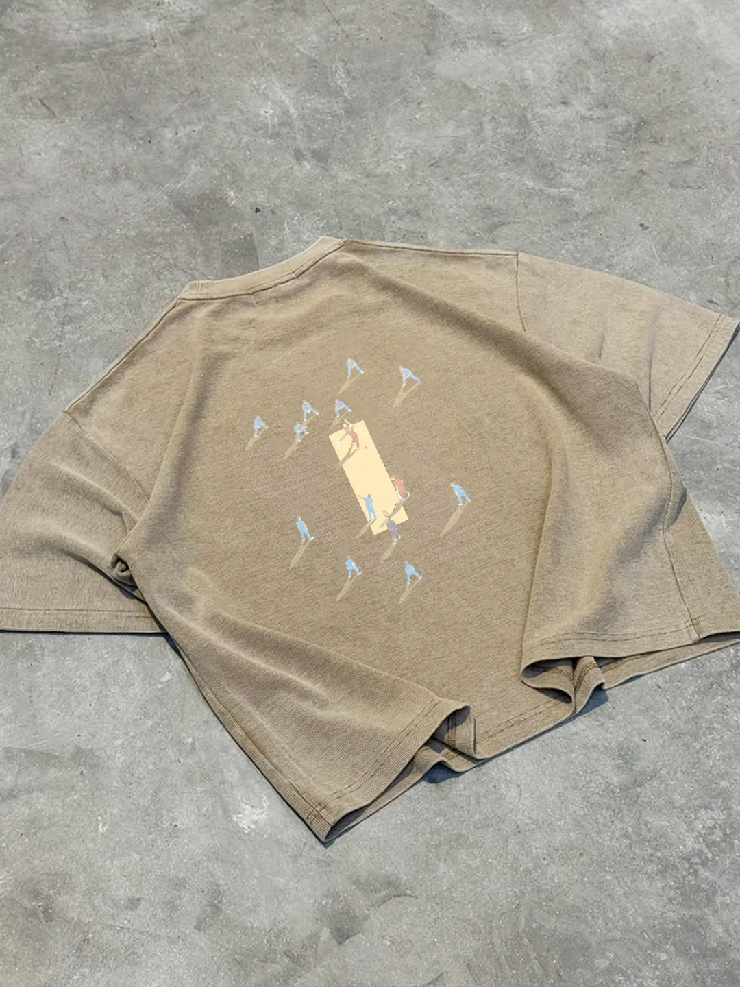 A beige short-sleeve t-shirt laid flat on a textured grey concrete floor, featuring a graphic print of a central beige rectangle surrounded by small blue and red figures.