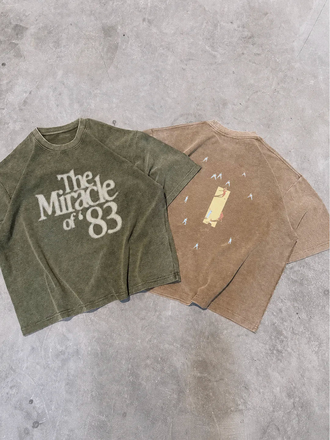 Two vintage-style short-sleeve t-shirts laid flat on a grey concrete floor. The t-shirt on the left is olive green with a distressed white graphic reading 'The Miracle of 83'.
