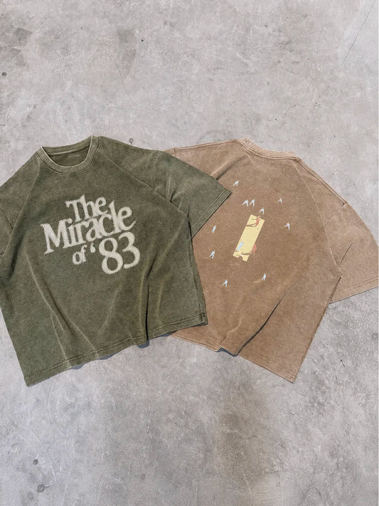 Two vintage-style short-sleeve t-shirts laid flat on a grey concrete floor. The t-shirt on the left is olive green with a distressed white graphic reading 'The Miracle of 83'.