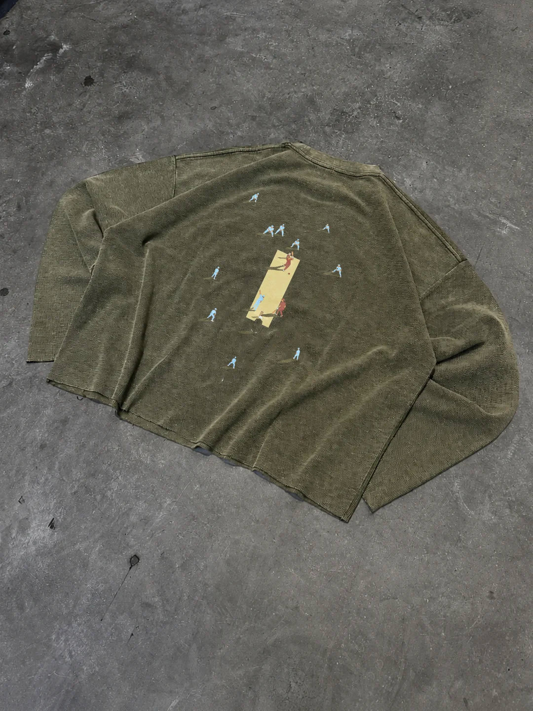 An olive green long-sleeve knit t-shirt with a central graphic print, laid flat on a textured grey concrete floor.