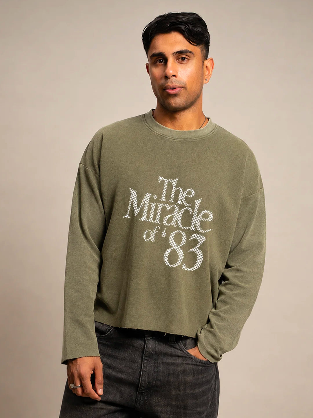 A man wearing an olive green long-sleeve crewneck t-shirt with a distressed white graphic reading 'The Miracle of '83' on the front, paired with dark grey jeans
